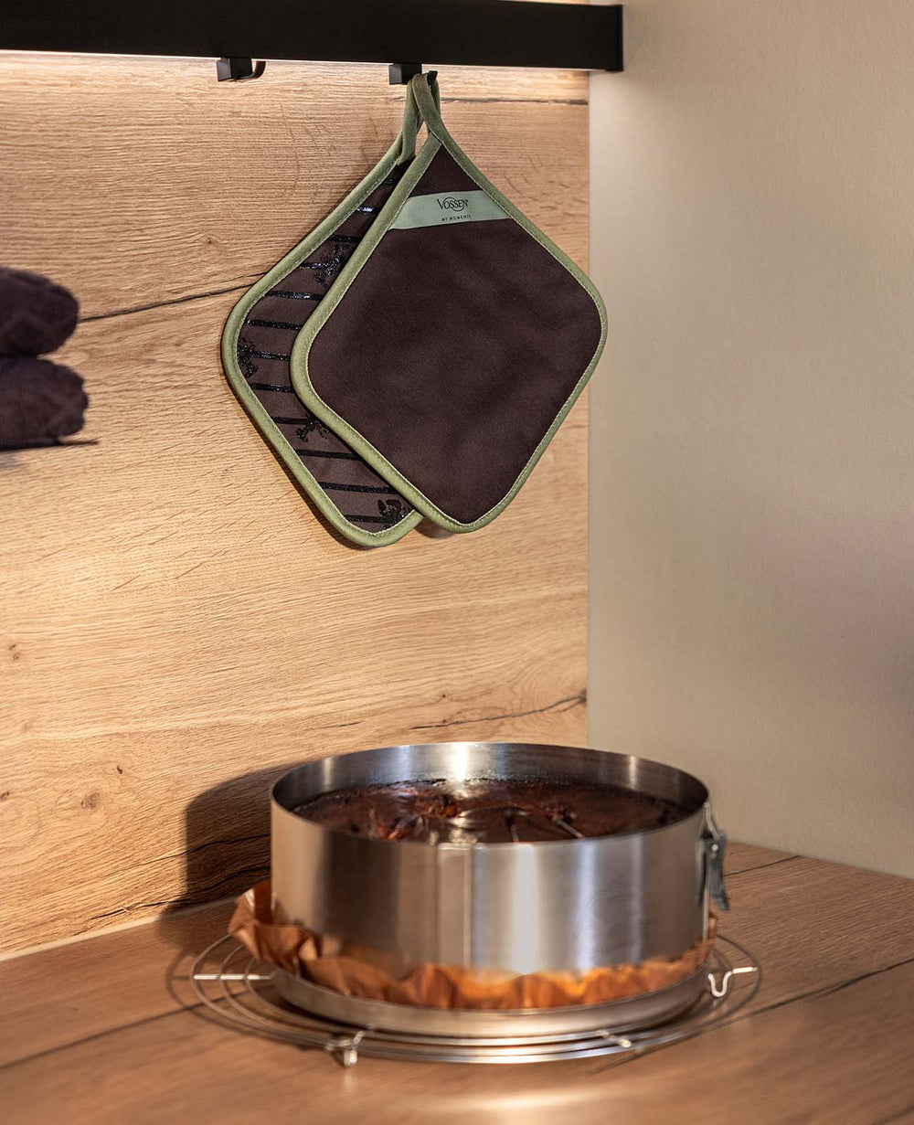 Zwei braune VOSSEN Topflappen mit olivgrüner Einfassung hängen an einem Haken vor einer warm beleuchteten Holzwand. Darunter steht ein frisch gebackener Schokoladenkuchen in einer Springform auf einem Kuchengitter.