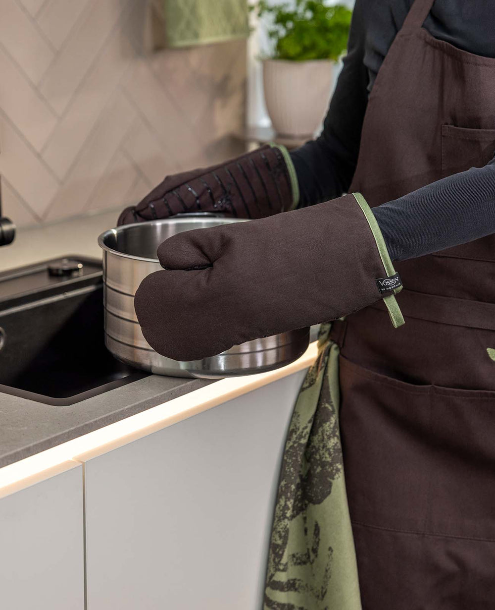 Eine Person mit dunkelbrauner Küchenschürze und passenden VOSSEN Ofenhandschuhen mit olivgrüner Einfassung hält einen Edelstahltopf in einer modernen Küche. Ein grün gemustertes Küchentuch ergänzt das praktische Küchenoutfit.