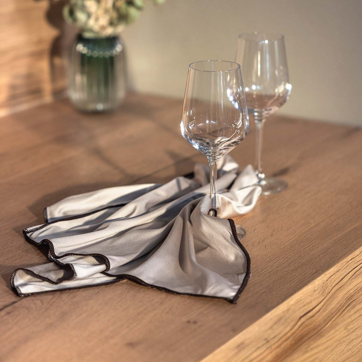 Zwei elegante Weingläser auf einem Holztisch, dekorativ arrangiert auf einem hellbeigen Tuch mit dunkler Einfassung. Warmes Sonnenlicht sorgt für eine gemütliche Atmosphäre.