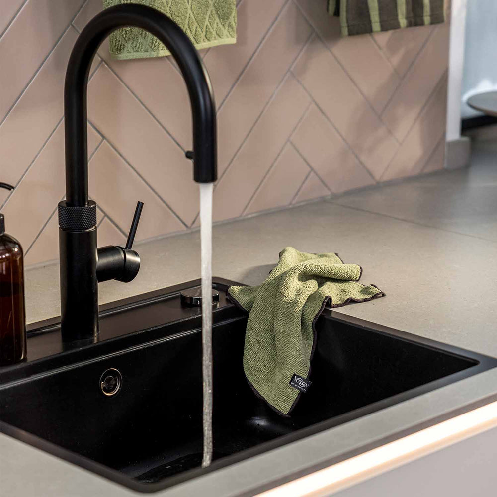 Grünes VOSSEN Küchentuch aus strukturierter Baumwolle, lässig über den Rand einer schwarzen Küchenspüle drapiert. Eine moderne schwarze Armatur mit fließendem Wasser und helle Fliesen unterstreichen das stilvolle Küchenambiente.
