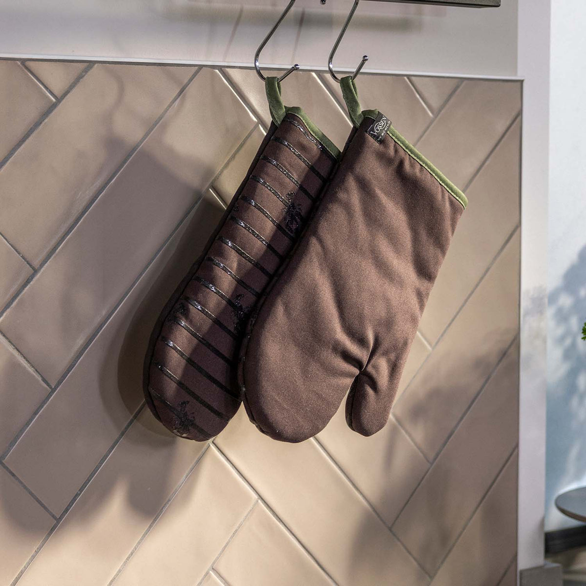 Zwei braune VOSSEN Ofenhandschuhe mit olivgrüner Einfassung hängen an Metallhaken vor einer hell gefliesten Küchenwand. Ein Handschuh ist einfarbig, der andere mit einem dezenten Streifenmuster gestaltet.