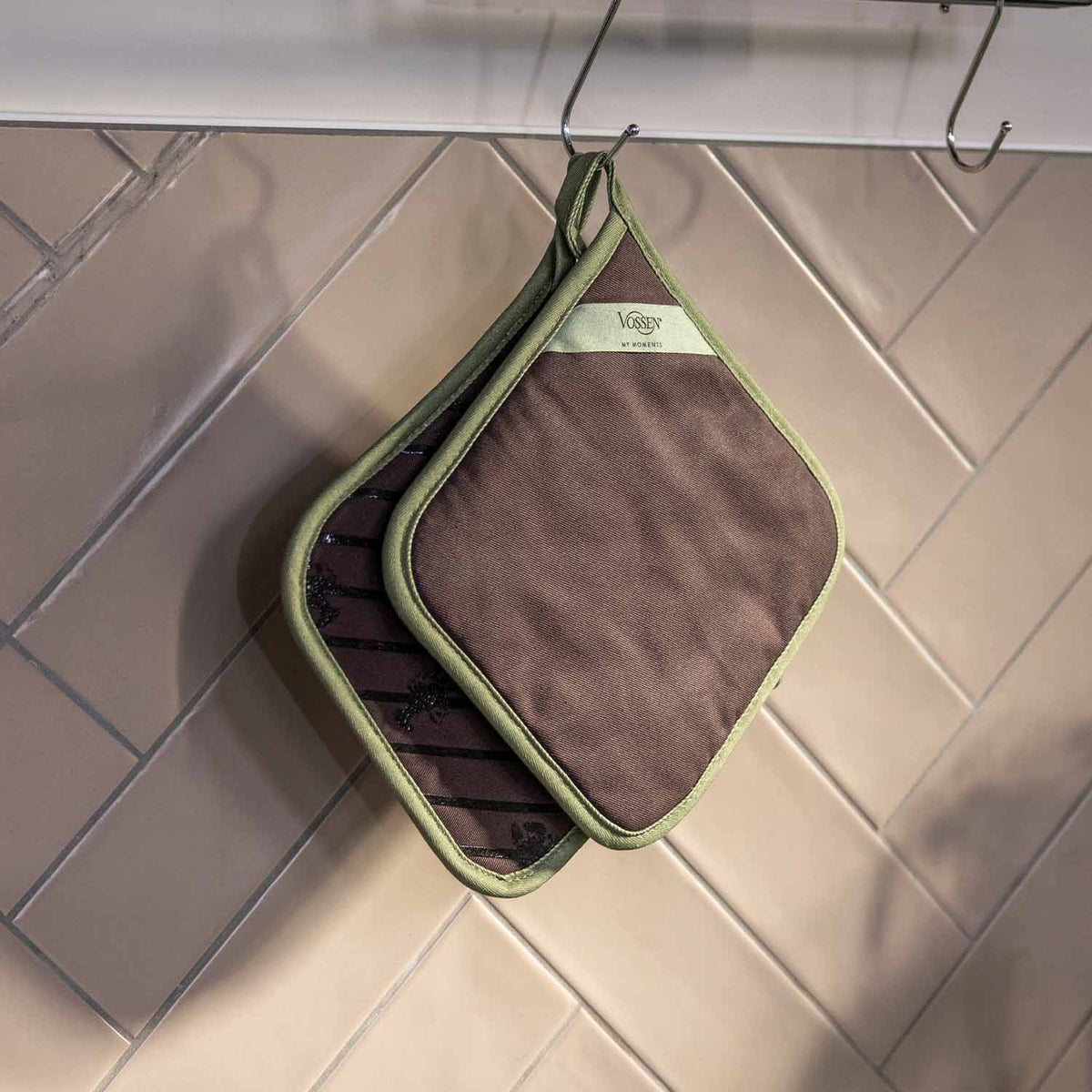 Zwei braune VOSSEN Topflappen mit grüner Einfassung, platzsparend an Metallhaken vor einer hell gefliesten Küchenwand aufgehängt.