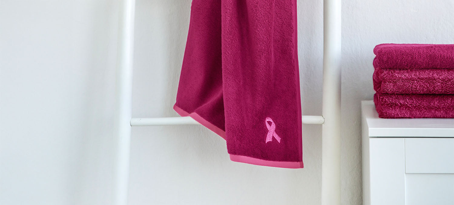Ein pinkfarbenes Handtuch mit einer aufgestickten Pink Ribbon-Schleife hängt an einer weißen Handtuchleiter. Im Hintergrund liegen weitere gefaltete Handtücher derselben Serie auf einem weißen Badmöbel