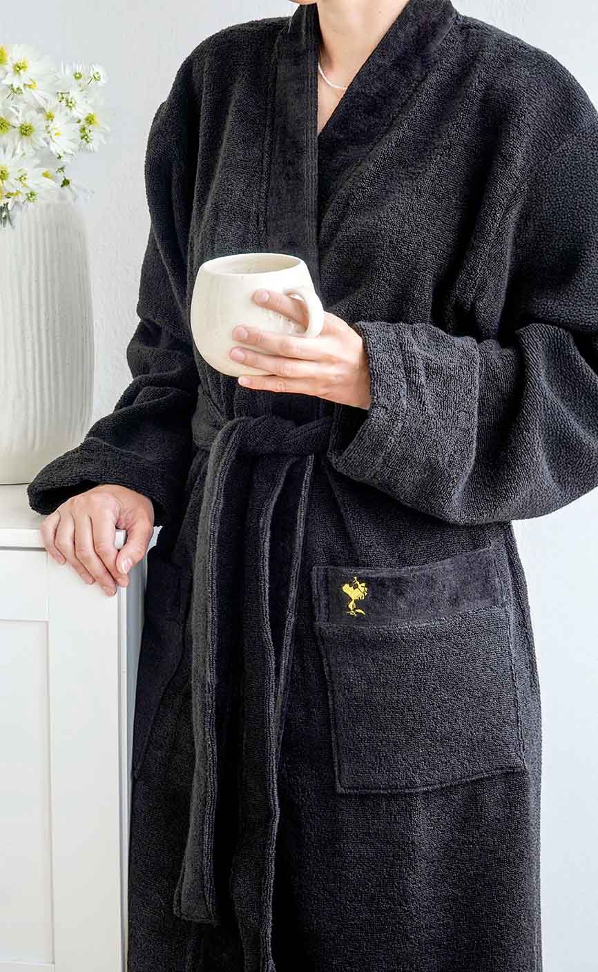 Person in schwarzem Bademantel mit gelber Woodstock-Stickerei hält eine helle Tasse – stilvolle Kombination aus Komfort und Peanuts-Design.