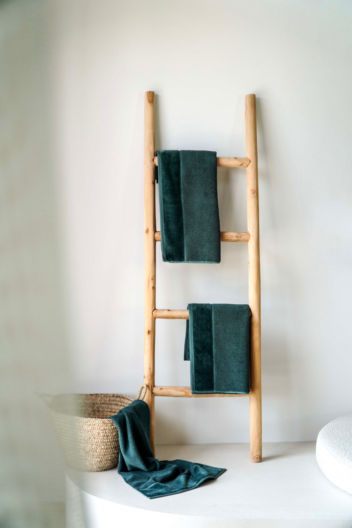 Dunkelgrüne VOSSEN Handtücher hängen dekorativ auf einer naturbelassenen Holzleiter vor einer hellen Wand. Ein geflochtener Korb mit einem weiteren Handtuch ergänzt das minimalistische Badezimmerambiente.