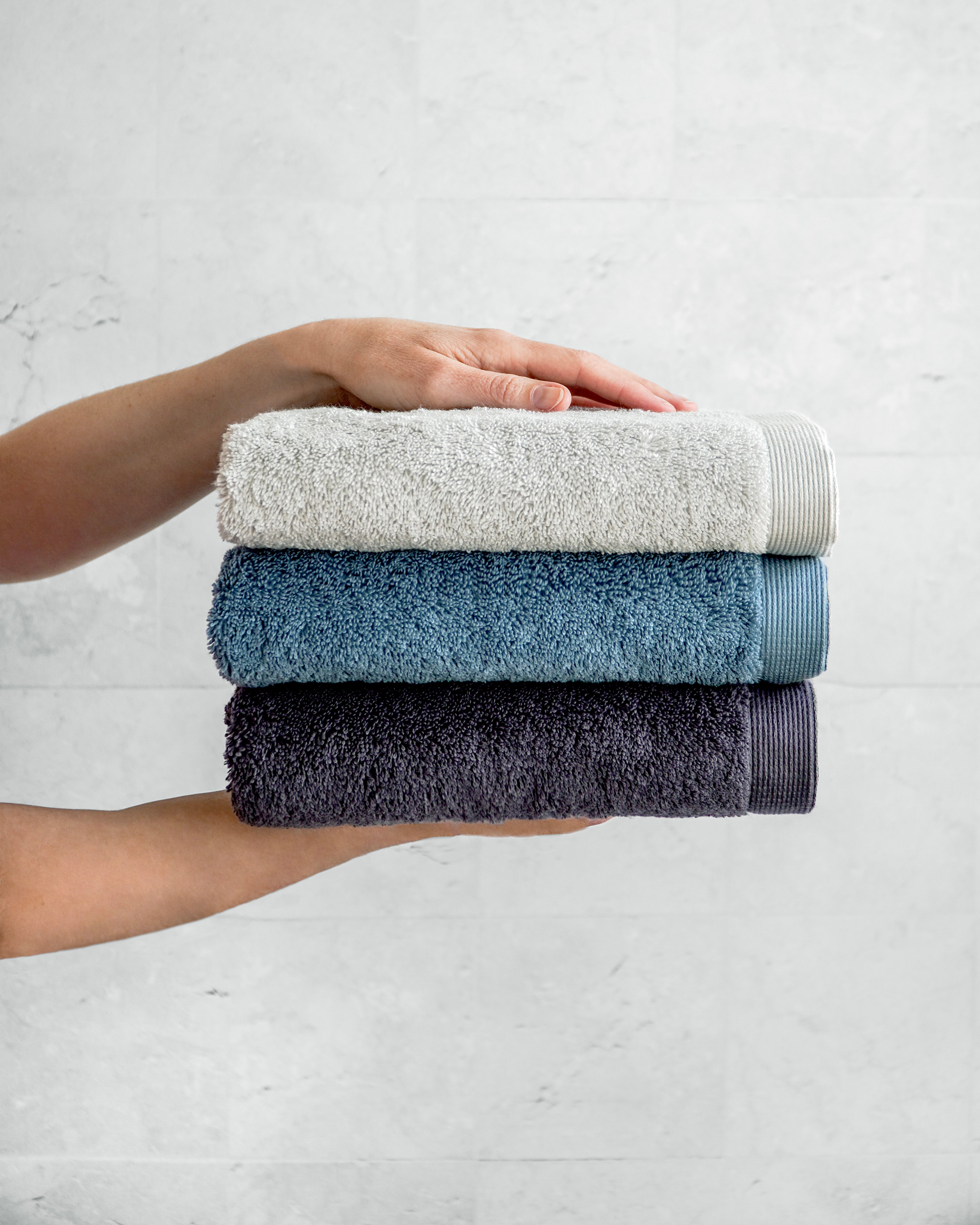 Hände halten drei gefaltete Handtücher in Grau- und Blautönen