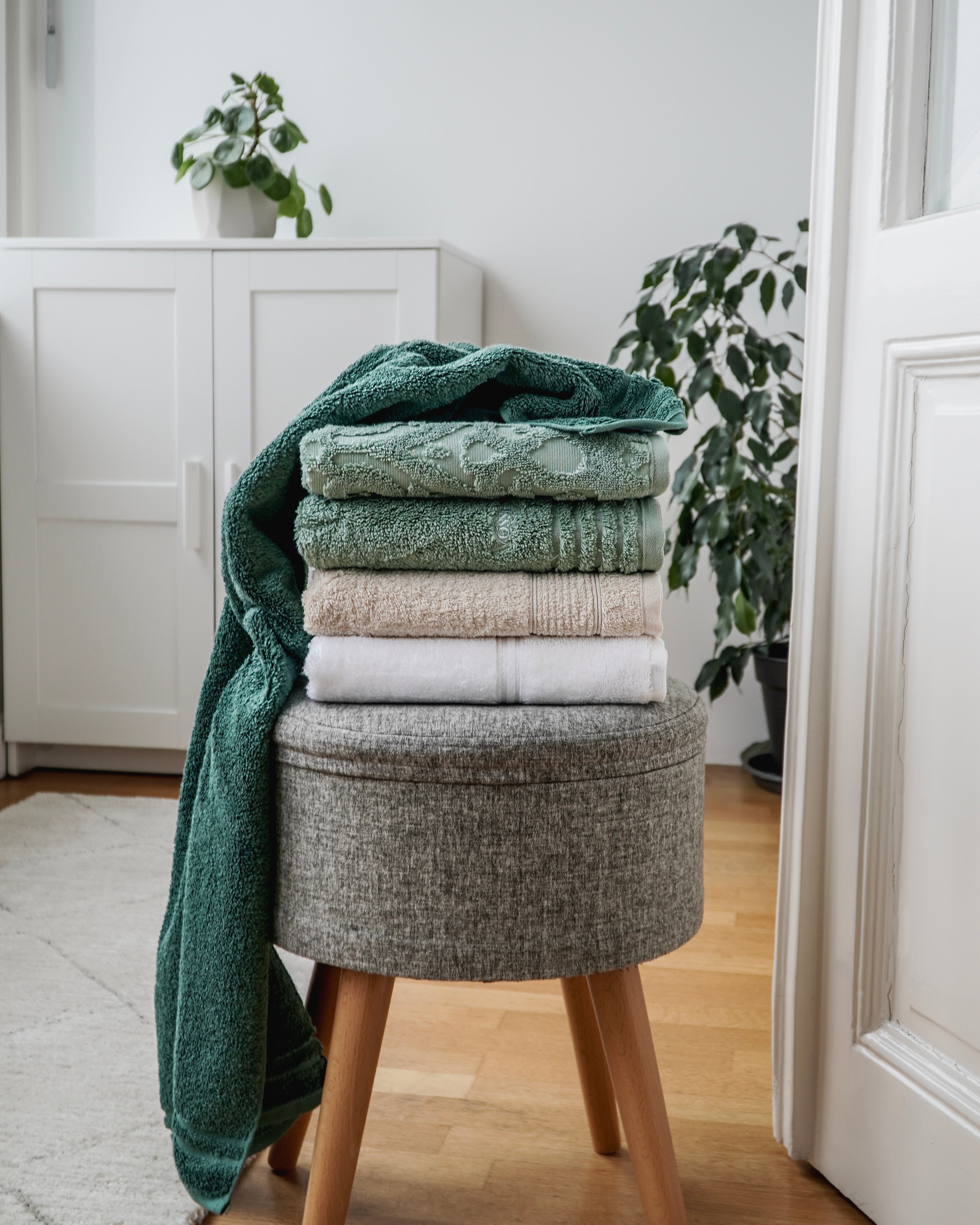 Gestapelte Handtücher in Grün- und Naturtönen auf einem Hocker im Wohnraum
