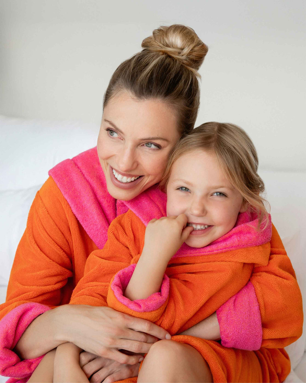 Eine lächelnde Mutter umarmt ihre Tochter auf einem hellen Sofa. Beide tragen kuschelige orangefarbene VOSSEN Bademäntel mit leuchtend pinkfarbenen Akzenten und genießen einen liebevollen Wohlfühlmoment.